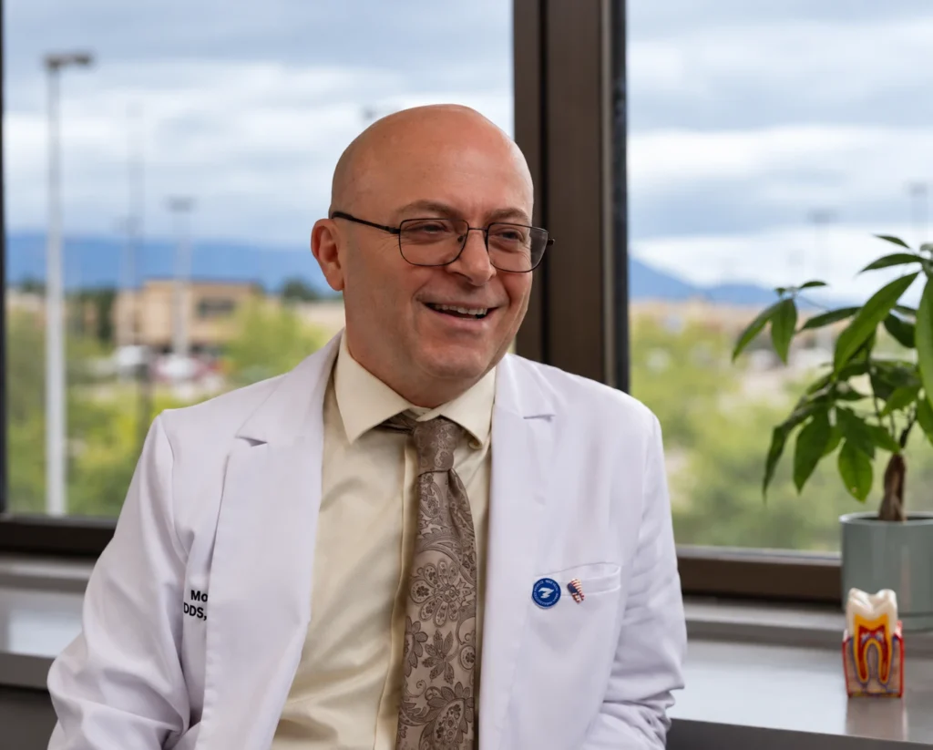 Smiling bald man in glasses wearing a white lab coat and patterned tie, seated by a large window with greenery outside; calm, welcoming mood.
