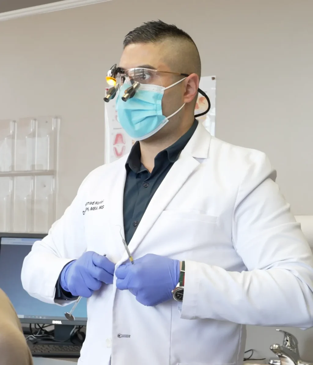 Dr Hariri, a dental professional in a white coat, protective goggles, and blue gloves, prepares an instrument beside a patient in a bright clinic, conveying care.